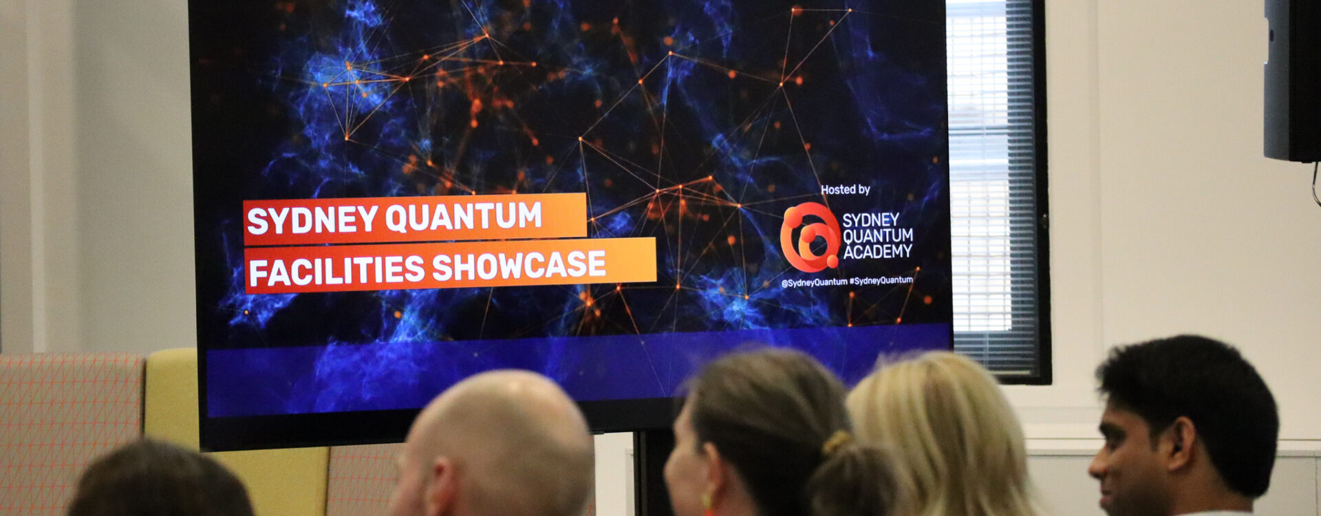 tv screen with words Sydney Quantum Facilities showcase in focus with blurred back of seated attendees