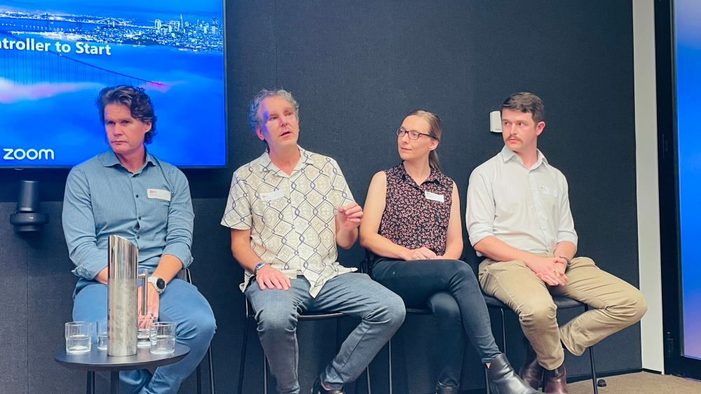 A group of four people - three men and one woman (second from right) - sit in front of a projector screen as part of a panel event.