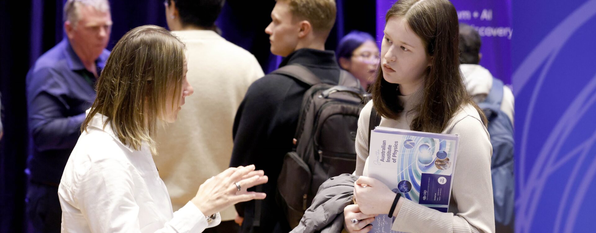 Two women stand in discussion in front of banners at a careers expo. A group of three men are in the crowd behind them.