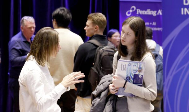 Two women stand in discussion in front of banners at a careers expo. A group of three men are in the crowd behind them.