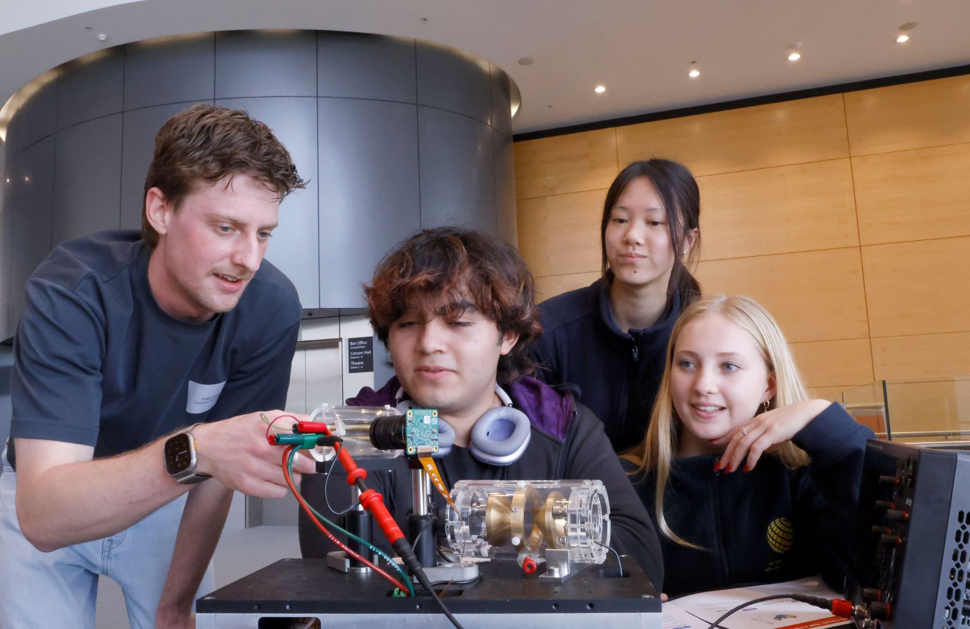 A group of high school students inspect a quantum sensor with a man at an expo booth